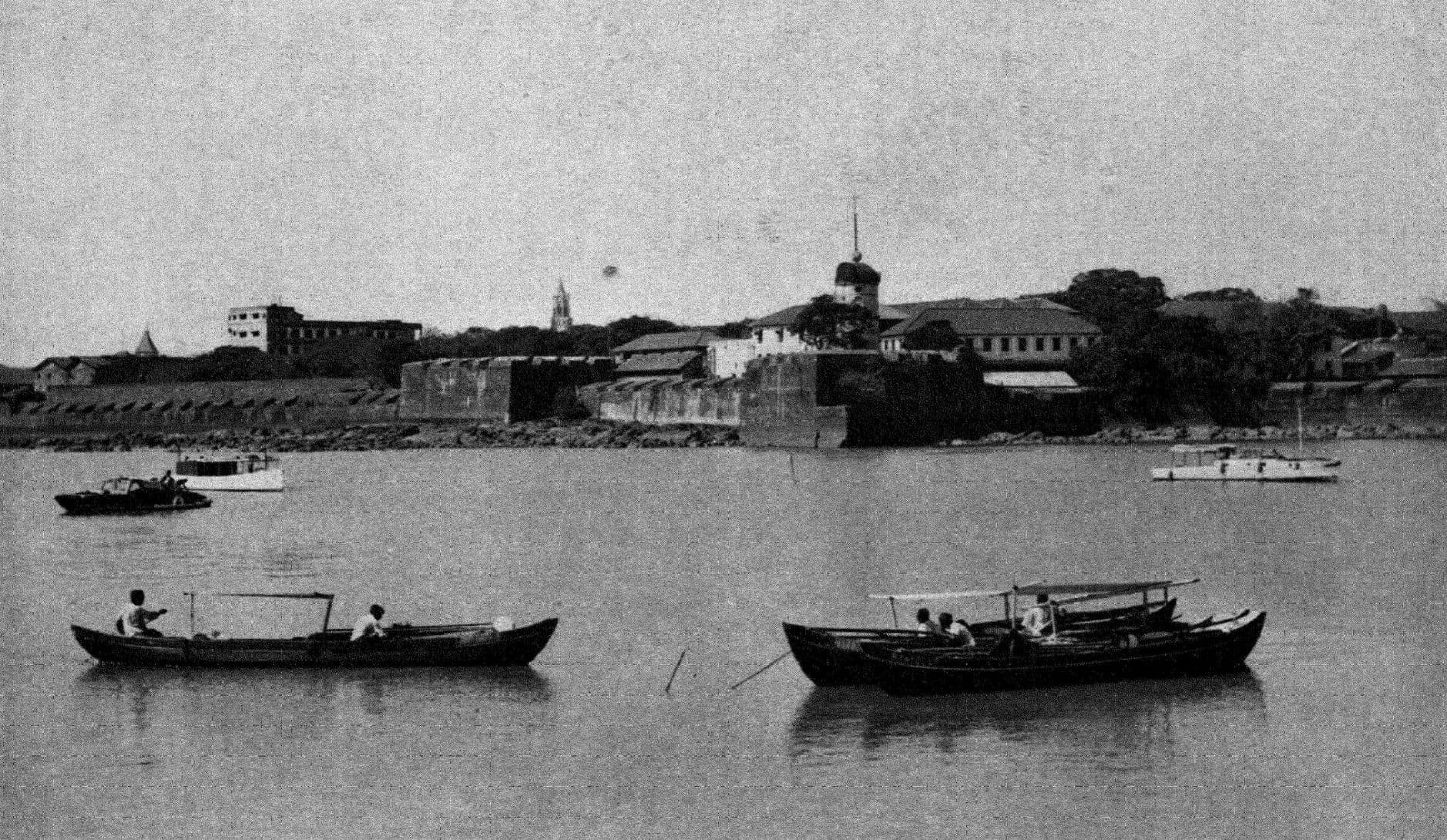 Rowing boats on calm water in front of the old walls of Bombay Fort and the buildings behind them.