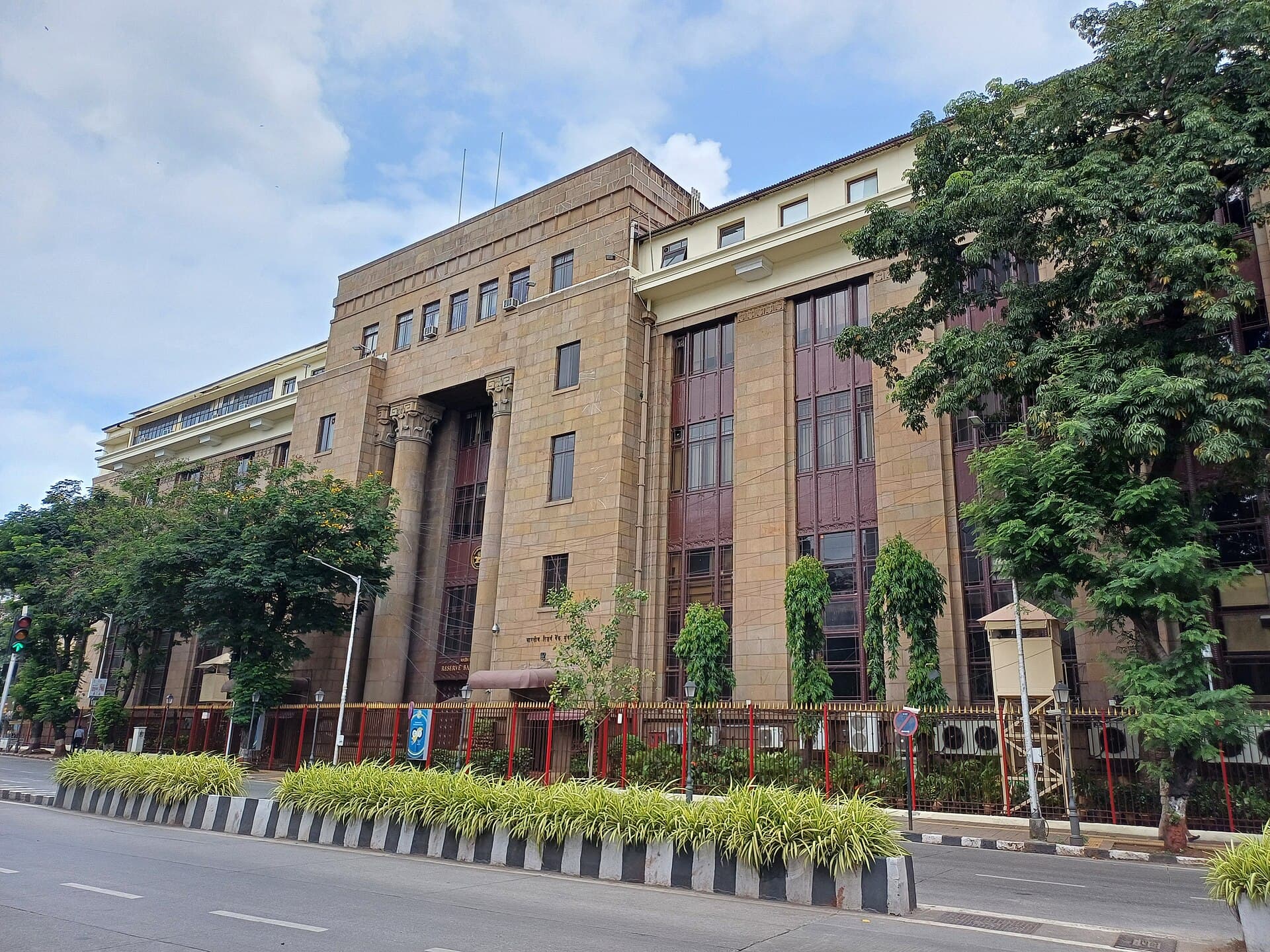 The stone façade and tall columns of the Reserve Bank of India building in Mumbai, behind a tree-lined street.